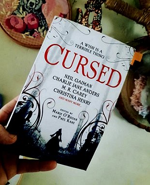 A hand holding a copy of the anthology Cursed, edited by Marie O'Regan and Paul Kane