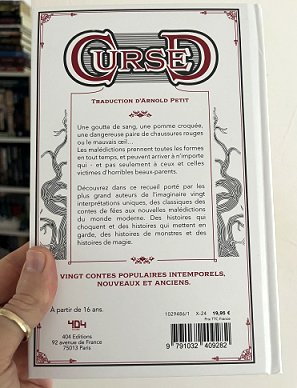 photograph of a man's hand holding up a copy of the French edition of Cursed, edited by Marie O'Regan and Paul Kane, to show the back cover