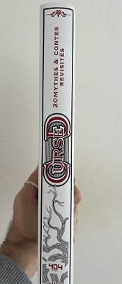 photograph of a man's hand holding up a copy of the French edition of Cursed, edited by Marie O'Regan and Paul Kane, to show the spine of the book