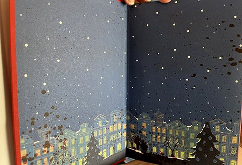 photograph showing the beautiful endpapers inside Death Comes at Christmas, edited by Marie O'Regan and Paul Kane. The endpapers feature a row of houses with lit up windows, Christmas trees and garlands, with snow falling in a grey sky. To the left, there's blood spatter