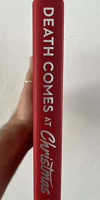 photograph showing a man's arm holding up a copy of Death Comes at Christmas, edited by Marie O'Regan and Paul Kane, against a white background to show the red bookjacket and silver text on the spine