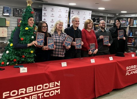 Death Comes at Christmas authors and editors standing behind a long table decorate with red tablecloths bearing the Forbidden Planet logo. All are holding up a copy of Death Comes at Christmas, edited by Marie O'Regan and Paul Kane. L to R: Tina baker (dressed as a Christmas tree), Fiona Cummins, Helen Fields, Paul Kane, Marie O'Regan, Tom Mead and Russ Thomas (dressed as Death)