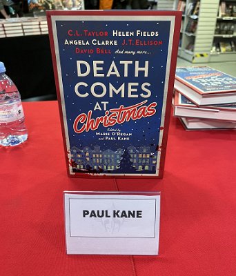 photo of Death Comes at Christmas, edited by Marie O'Regan and Paul Kane, standing on a red tablecloth behind a nameplate showing the name PAUL KANE