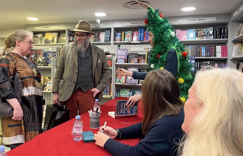 Tina Baker (dressed as a Christmas tree) signing a copy of Death Comes at Christmas, edited by Marie O'Regan and Paul Kane, for two customers. In the foreground is Helen Fields, with Fiona Cummins behinid her