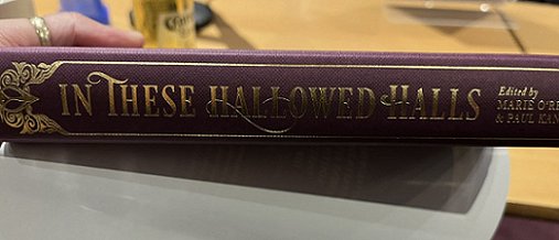 A hand with a gold thum bring holds a copy of In These Hallowed Halls, edited by Marie O'Regan and Paul Kane, to display the gold lettering on the spine of the book