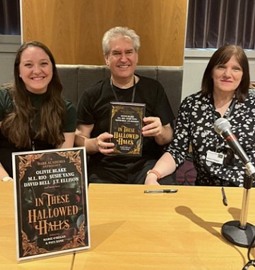 L to R: Tori Bovalino, Paul Kane and Marie O'Regan sit at a table behind a poster of In These Hallowed Halls, edited by Marie O'Regan and Paul Kane. Paul is holding up a copy of the book