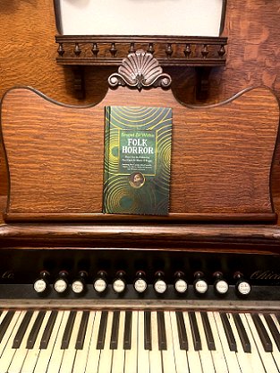 photograph of a copy of Beyond and Within Folk Horror, edited by Paul Kane and Marie O'Regan, on the music shelf of a wooden organ