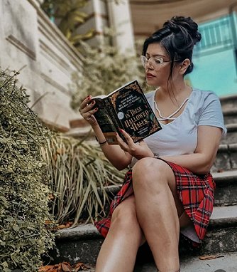 photograph of a young woman with dark hair piled up into a bun, wearing glasses and a white t shirt, red plaid skirt, sitting on steps and reading a copy of In These Hallowed Halls, edited by Marie O'Regan and Paul Kane