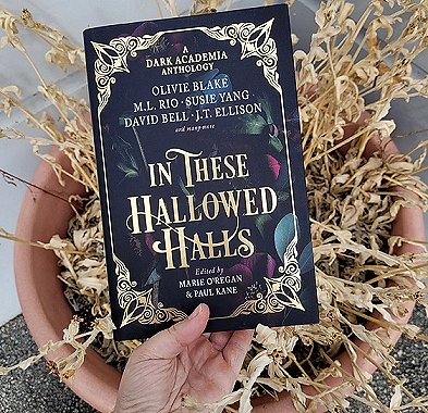 A hand holds a copy of In These Hallowed Halls, edited by Marie O'Regan and Paul Kane, above a terracotta planter with dying plant inside