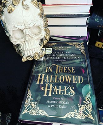 image showing a copy of In These Hallowed Halls, edited by Marie O'Regan and Paul Kane, lying on a black cloth in front of a pile of hardback books. A white skull ornament decorated with gold roses is off to the lefthand side at the top of the book