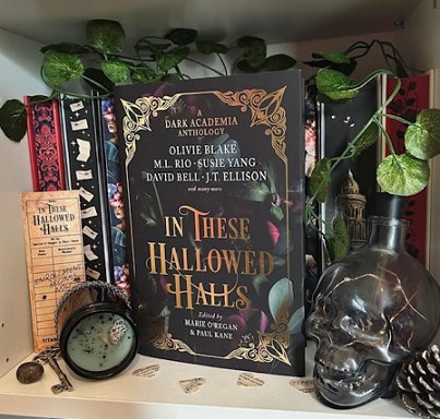 image showing a copy of In These Hallowed Halls standing on a white shelf in front of a row of books showing their sprayed edges. To the left of the book is a bookmark for In These Hallowed Halls, alongside a key and a pot with a clear glass lid. To the right is a black skull holding a black candle and a pine cone. Ivy leaves twist along the tops of the shelf above the books