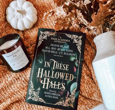 image showing a copy of In These Hallowed Halls, edited by Marie O'Regan and Paul Kane, lying on an orange knitted throw, beside a candle in a brown glass jar, a white pumpkin ornament and a white vase with autumnal leaves inside