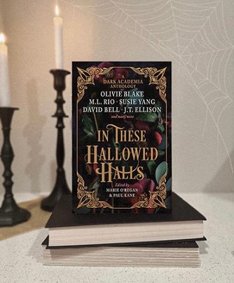 Image showing a copy of In These Hallowed Halls, edited by Marie O'Regan and Paul Kane, standing on top of two closed black books on a cream surface with two black candelabras and lit white candles bedhind them, and a spiderweb in the righthand top corner