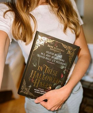 photograph of a woman with long blonde hair, wearing a white t shirt and blue jeans, holding a copy of In These Hallowed Halls, edited by Marie O'Regan and Paul Kane