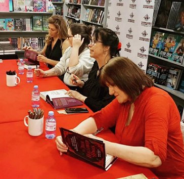 authors signing copies of In These Hallowed Halls, edited by Marie O'Regan and Paul Kane, seated at a table with a red cloth covering it and pots of pens on the table alongside bottles of water. L to R: Kate Weinberg, Tori Bovalino, Helen Grant and Marie O'Regan