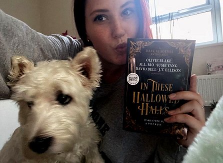 Image of a woman sitting down with a white dog, holding up a copy of In These Hallowed Halls, edited by Marie O'Regan and Paul Kane