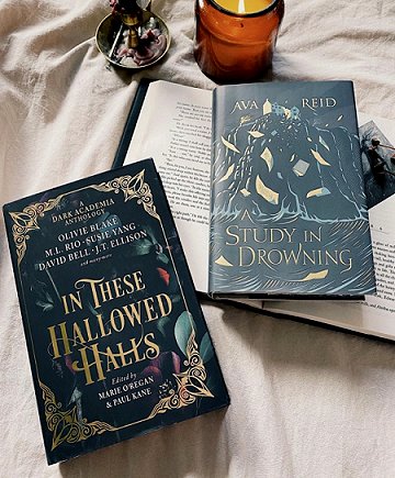 photograph showing a copy of In These Hallowed Halls, edited by Marie O'Regan and Paul Kane, and a copy of A Study in Drowning by Ava Reid lying on an open book on a beige cloth. At the top of the picture are a lit jar candle and a brass candle holder