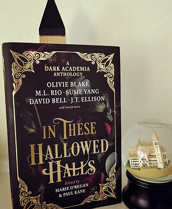 photograph of a copy of In These Hallowed Halls, edited by Marie O'Regan and Paul Kane, standing in front of a white background. To the righthand side is a glass dome with a model castle inside, on a dark wooden plinth