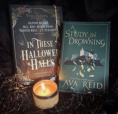 Two books propped on dark shredded tissue paper behind a lit jar candle. In These Hallowed Halls, edited by Marie O'Regan and Paul Kane, and A Study in Drowning by Ava Reid