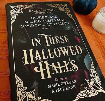A copy of In These Hallowed Halls, edited by Marie O'Regan and Paul Kane, lying on a wooden surface - a jar candle is alongside the book