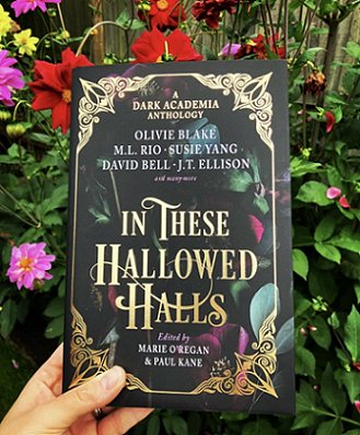 a hand holds a copy of In These Hallowed Halls, edited by Marie O'Regan and Paul Kane, in front of flowering bushes