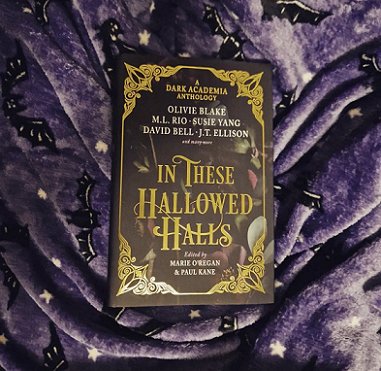 A copy of In These Hallowed Halls, edited by Marie O'Regan and Paul Kane, lies on tp of a purple fleece cloth decorated with black cats and white stars