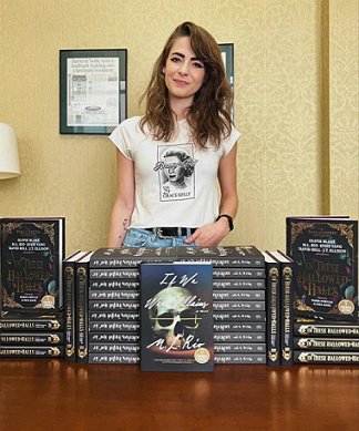 photograph showing a smiling M L Rio standing behind a stack of her novel, If We Were Villains, and a stack of In These Hallowed Halls, edited by Marie O'Regan and Paul Kane
