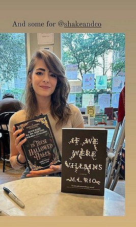photograph of M L Rio sitting at a table in a bookstore behind a standing copy of If We Were Villains, holding a copy of In These Hallowed Halls, edited by Marie O'Regan and Paul Kane