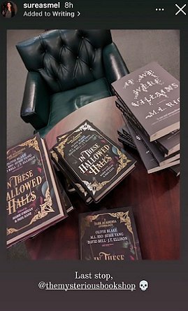 photograph of a round wooden table with copies of If We Were Villains by M L Rio alongside copies of In These Hallowed Halls, edited by Marie O'Regan and Paul Kane. A green leather chair sits behind the table