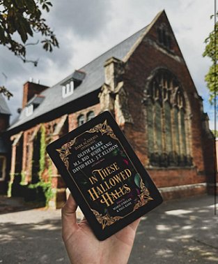 Image showing a hand holding a Kindle featuring In These Hallowed Halls, edited by Marie O'Regan and Paul Kane, against the background of an old church building