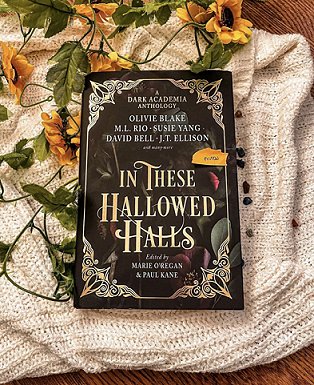 photograph of a copy of In These Hallowed Halls, edited by Marie O'Regan and Paul Kane, lying on a cream knitted cloth, with yellow flowers and green leaves underneath