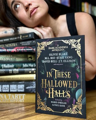 Olivie Blake leans on a stack of Dark Academia novels, in front of which stands a copy of In These Hallowed Halls, edited by Marie O'Regan and Paul Kane
