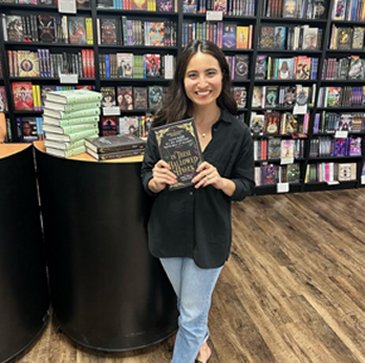 photograph of author Olivie Blake in a bookstore, standing in front of a display of her novels and full bookshelves, holding a copy of In These Hallowed Halls, edited by Marie O'Regan and Paul Kane