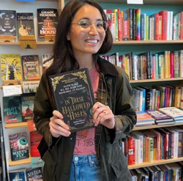photograph of a smiling author, Olivie Blake, standing in front of packed bookshelves, holding a copy of In These Hallowed Halls, edited by Marie O'Regan and Paul Kane