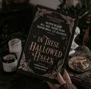 photograph showing a woman's hand holding a copy of In These Hallowed Halls, edited by Marie O'Regan and Paul Kane, above a brown cloth on which stand a jar candle and a coaster