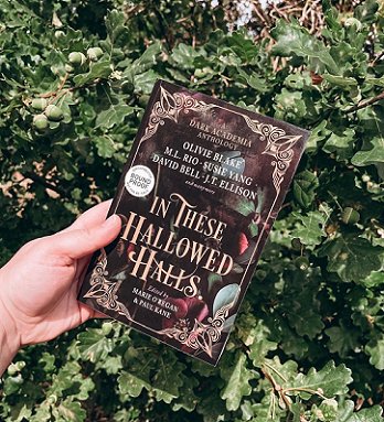Image of a hand holding a copy of In These Hallowed Halls, edited by Marie O'Regan and Paul Kane, against a background of green leaves and acorns