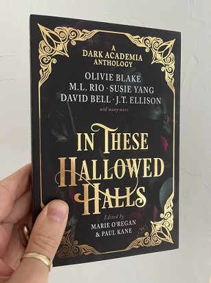 photograph of a man's hand holding a paperback copy of In These Hallowed Halls, edited by Marie O'Regan and Paul Kane, up against a white background