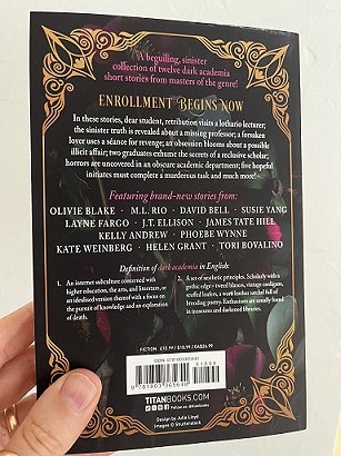 photograph of a man's hand holding up a copy of In These Hallowed Halls, edited by Marie O'Regan and Paul Kane, to show the back cover against a white background