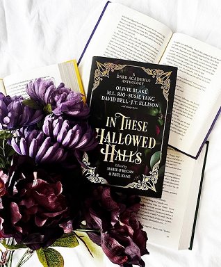 image showing a copy of In These Hallowed Halls, edited by Marie O'Regan and Paul Kane, lying on three open books on a white surface. To the left is a bouquet of purple and dark red flowers with foliage
