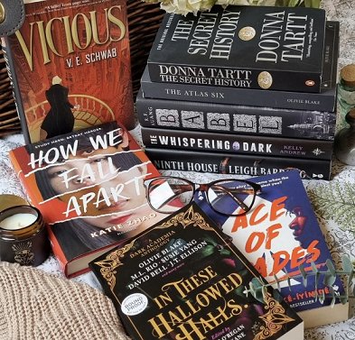 photograph showing a pile of Dark Academia novels on a white cloth decorated with green leaves, and a proof copy of In These Hallowed Halls, edited by Marie O'Regan and Paul Kane, in the front on a beige knitted cloth. A black-framed pair of glasses lie on top, and there's a candle in a brown glass jar