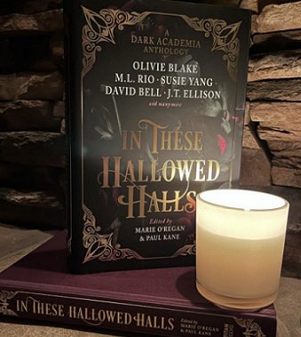 image showing a copy of In These Hallowed Halls, edited by Marie O'Regan and Paul Kane, standing on top of a copy of the book that has no dustjacket, revealing its purple cover and the gold text on the spine. Standing on top of this is a votive candle