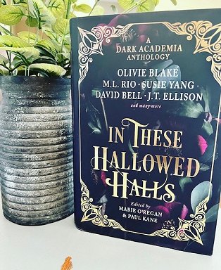 image of a copy of In These Hallowed Halls, edited by Marie O'Regan and Paul Kane, standing on a white surface. To its left is a grey planter, with green leaves growing from the top