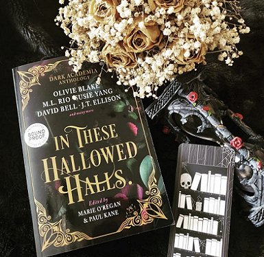 photograph of a proof copy of In These Hallowed Halls, edited by Marie O'Regan and Paul Kane, lying on a back surface, beside a black bookmark decorated with white skulls and books. Above is a bouquet of cream roses and gypsophila, on a black and pink floral cloth