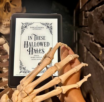 photograph of a hand holding a Kindle featuring the title page of In These Hallowed Halls, edited by Marie O'Regan and Paul Kane. A skeleton is holding its hand over the hand holding the Kindle