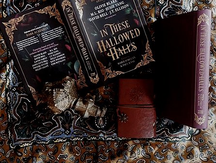 photograph featuring a copy of In These Hallowed Halls, edited by Marie O'Regan and Paul Kane, lying opn on a brown and purple paisley cloth, alongside a burgundy leather notebook and a copy of In These Hallowed Halls without a dustjacket, revealing the purple cover and gold lettering on the spine
