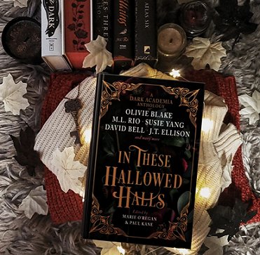 Image showing a copy of In These Hallowed Halls, edited by Marie O'Regan and Paul Kane, lying on a cream and red knitted background, with cream paper leaves and a key displayed alongside, and a candle and some hardback books above