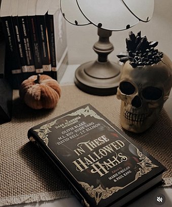 a copy of In These Hallowed Halls, edited by Marie O'Regan and Paul Kane, lies on a beige woven cloth beside a skull, a pumpkin ornament and a brass table lamp