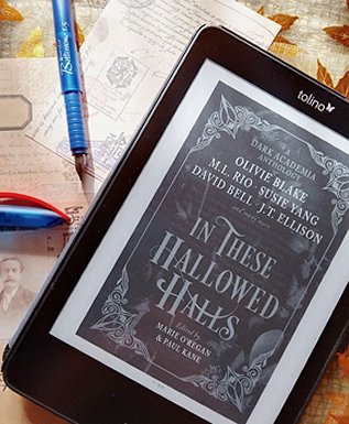 image shows a tablet reader, with the cover page of In These Hallowed Halls, edited by Marie O'Regan and Paul Kane, showing in black and white. The tablet is on a pile of old postcards, alongside a blue fountain pen with a blue and red lid