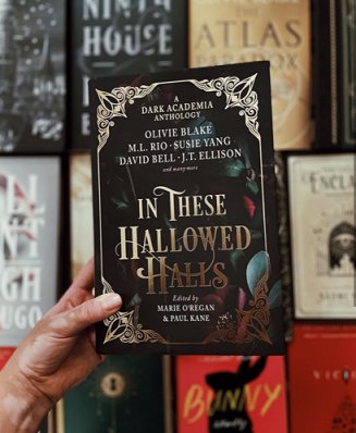 Image of a hand holding up a copy of In These Hallowed Halls, edited by Marie O'Regan and Paul Kane, against a background display of Dark Academia novels, including The Atlas Paradox by Olivie Blake, Ninth House by Leigh Bardugo