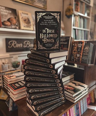 photograph of a spiral tower of copies of In These Hallowed Halls, edited by Marie O'Regan and Paul Kane, with one copy standing on top, at the front of a table displaying many other books in a bookstore